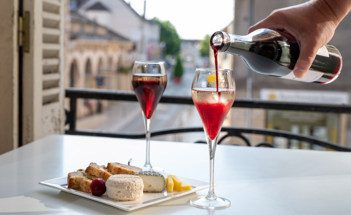 Aus einer großen Flasche wird rote Flüssigkeit in ein mit Weißwein oder Sekt gefülltes Glas gegossen, daneben ein bereits gemischtes Glas und ein Frühstück, auf einem Balkon